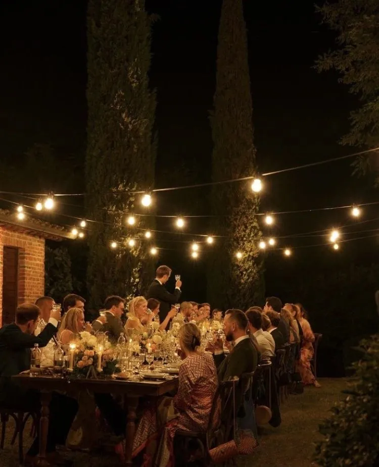 Cena de boda al aire libre entre cipreses, con invitados brindando bajo guirnaldas de bombillas