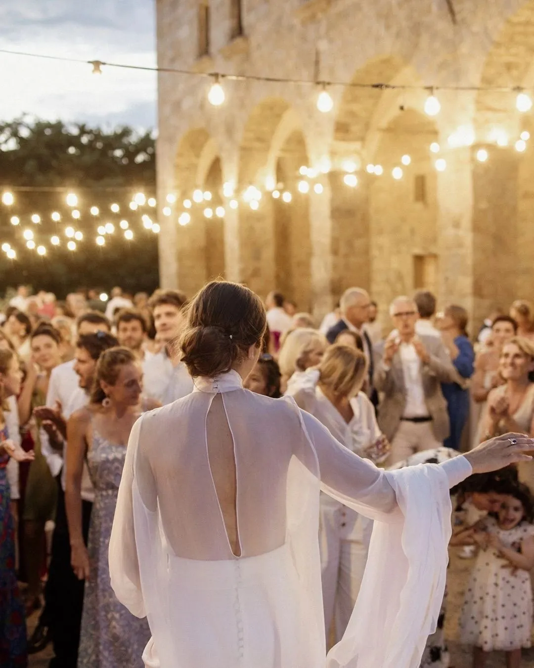 La novia de espaldas saludando a los invitados bajo guirnaldas de luces en un patio de piedra