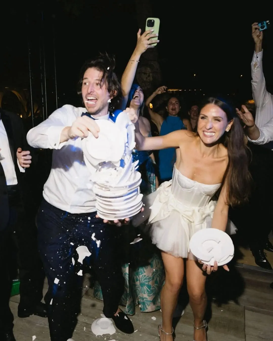Los novios rompiendo una torre de platos entre risas, con los invitados celebrando detrás